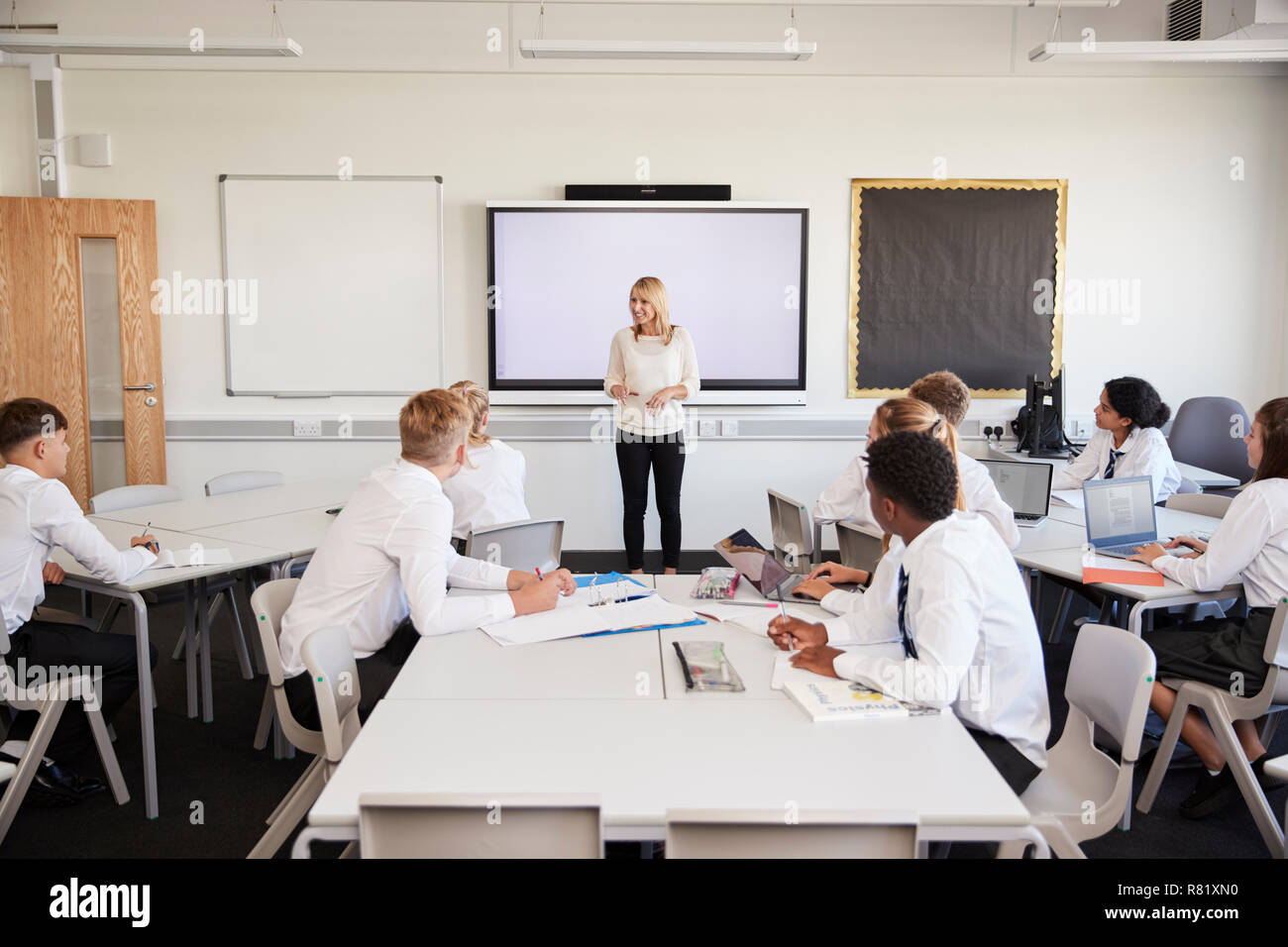Female High School Teacher Standing Next To Interactive Whiteboard And Teaching Lesson To Pupils Wearing Uniform Stock Photo