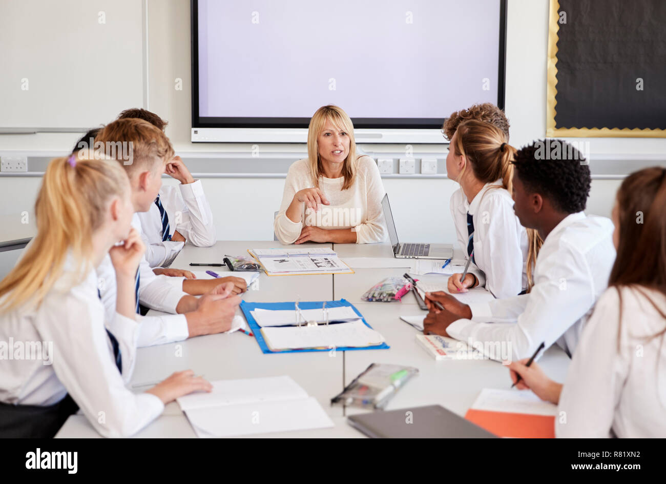 Female High School Teacher Sitting At Table With Teenage Pupils Wearing Uniform Teaching Lesson Stock Photo
