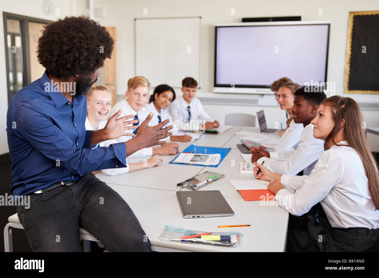 Male High School Teacher Sitting At Table With Teenage Pupils Wearing Uniform Teaching Lesson Stock Photo