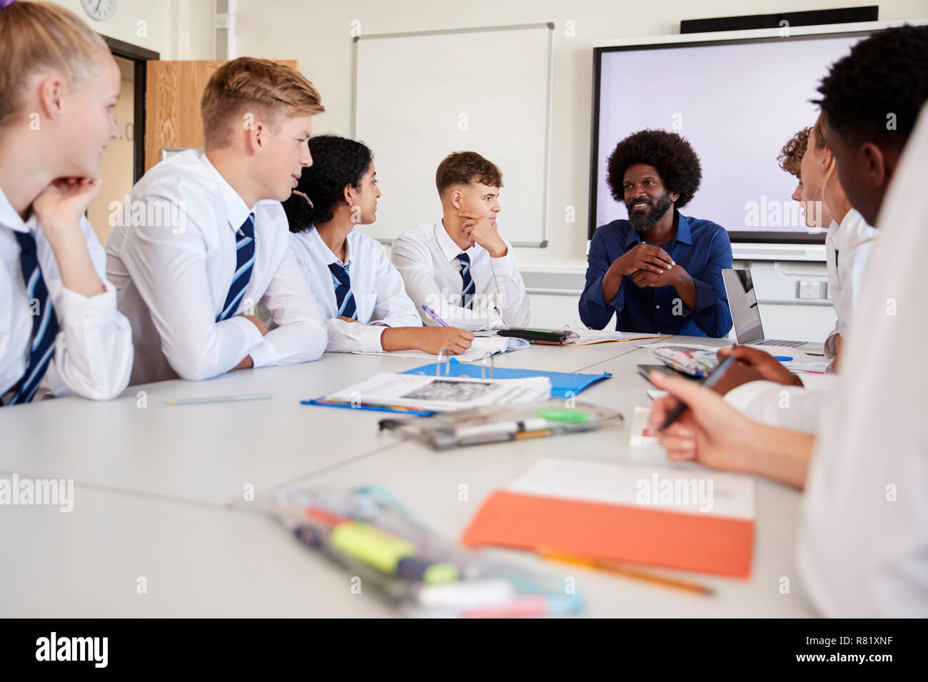 Male High School Teacher Sitting At Table With Teenage Pupils Wearing Uniform Teaching Lesson Stock Photo