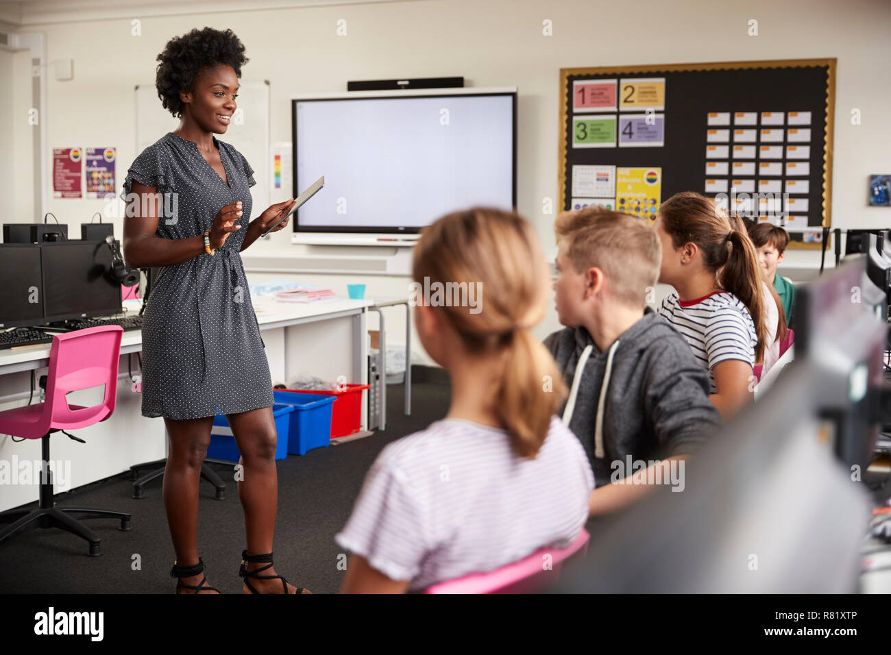 Teacher With Digital Tablet Talking To Line Of High School Students Sitting By Screens In Computer Class Stock Photo