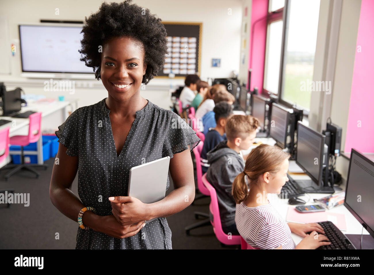 Portrait Of Female Teacher Holding Digital Tablet Teaching Line Of High School Students Sitting By Screens In Computer Class Stock Photo