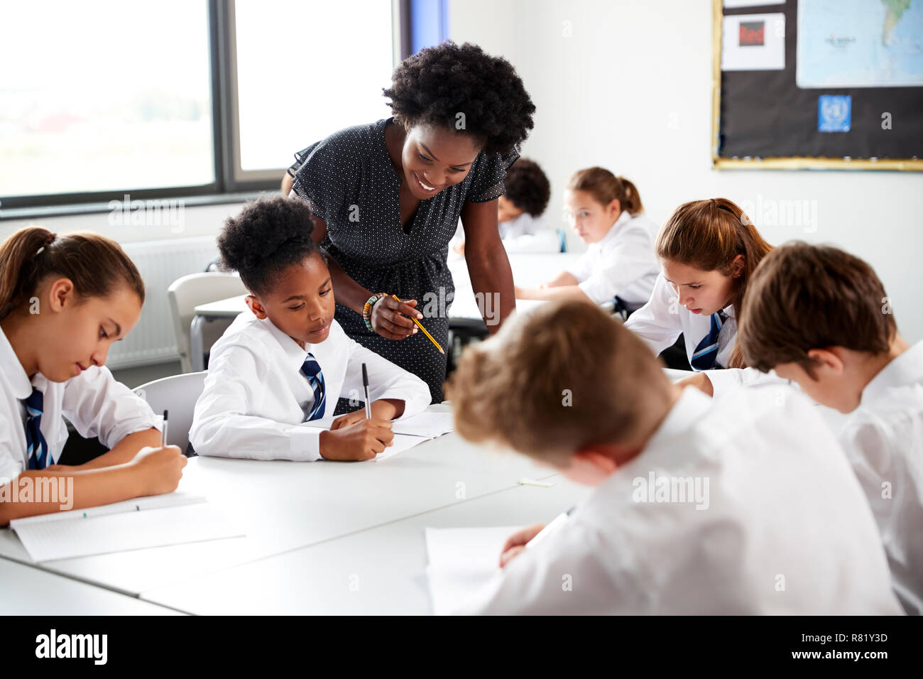 Female High School Tutor Helping Students Wearing Uniform Seated Around Tables In Lesson Stock Photo