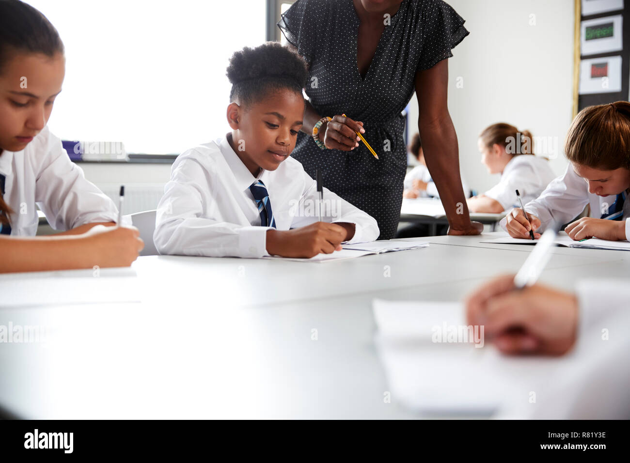 Female High School Tutor Helping Students Wearing Uniform Seated Around Tables In Lesson Stock Photo