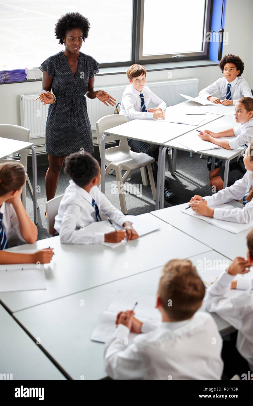 Female High School Tutor Helping Students Wearing Uniform Seated Around Tables In Lesson Stock Photo