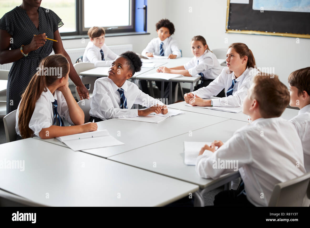 Detail Of Female High School Tutor Helping Students Wearing Uniform Seated Around Tables In Lesson Stock Photo