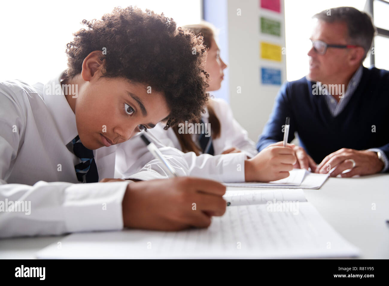 Male High School Student Wearing Uniform Working At Table With Teacher Talking To Pupils In Background Stock Photo
