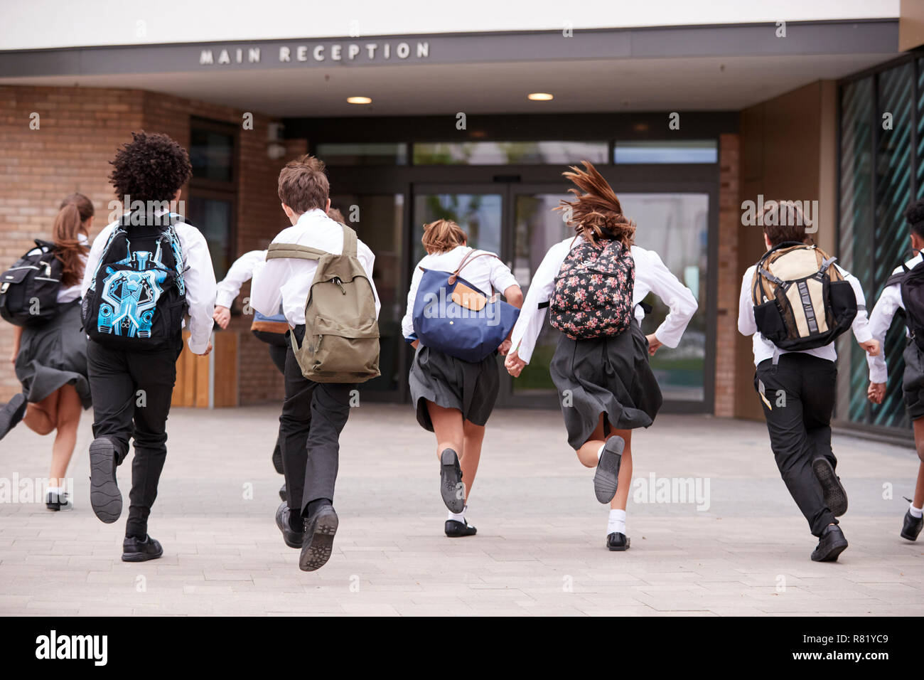 Group Of High School Students Wearing Uniform Running Into School Building At Beginning Of Class Stock Photo