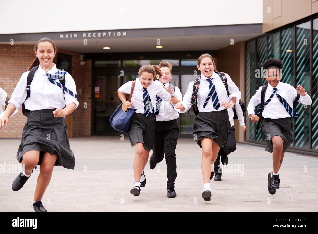 Group Of High School Students Wearing Uniform Running Out Of School Buildings Towards Camera At The End Of Class Stock Photo