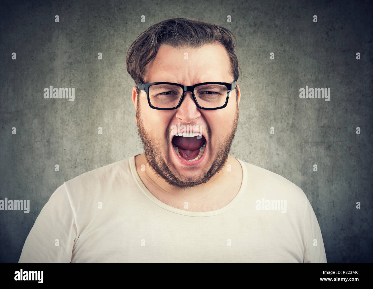 Young casual chubby man in glasses yelling at camera in anger on gray background Stock Photo