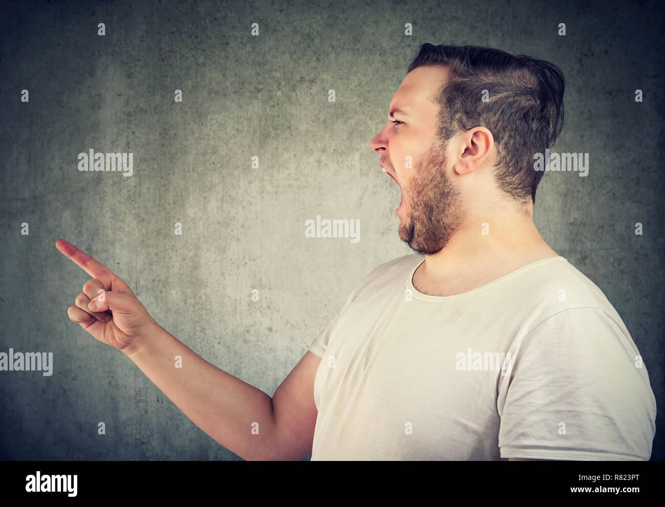 Young casual chubby man yelling in anger and pointing finger at someone on gray background Stock Photo