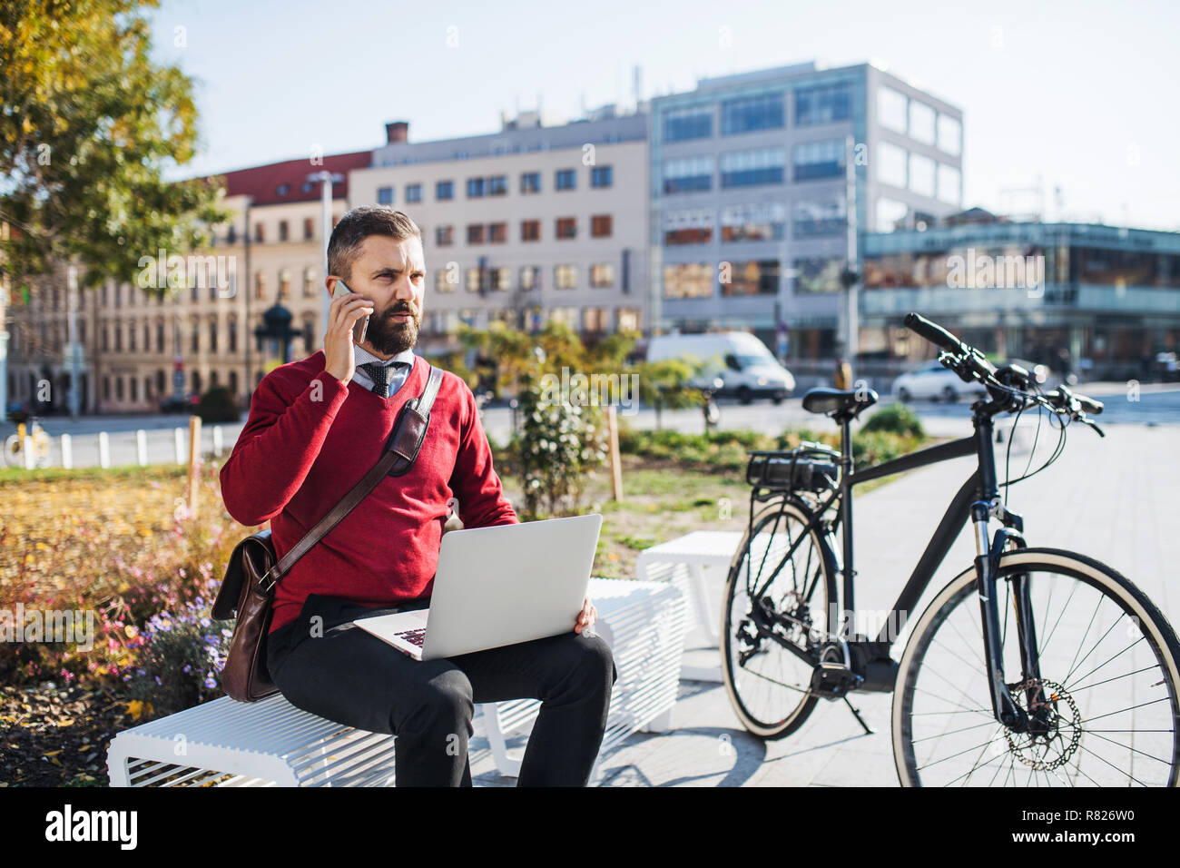 Businessman commuter with electric bicycle sitting on bench, using laptop and smartphone. Stock Photo