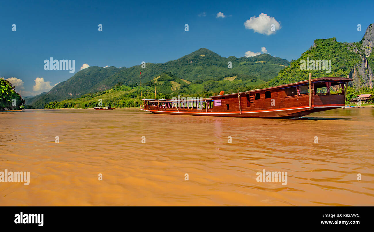 A long brown tour boat sails down the mighty Mekong River in Laos with the lush green mountains lining the shore Stock Photo