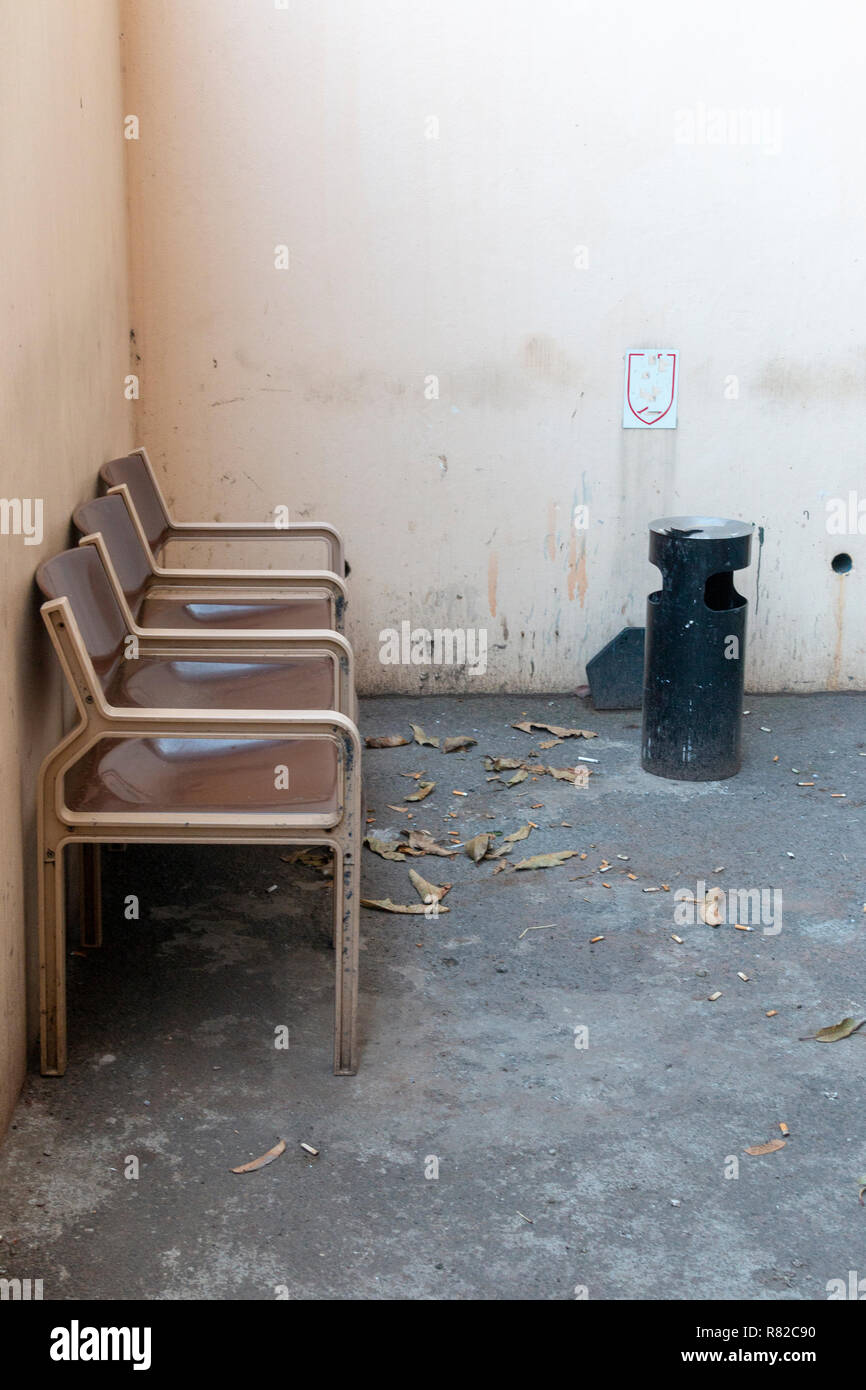 A close up front view of three brown and cream connected seats outside in a designated smoking area with a black ashtray bin in the conner Stock Photo