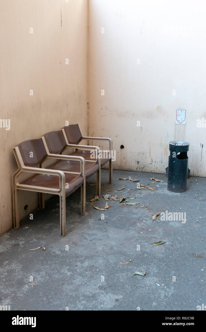 A close up side view of three brown and cream connected seats outside in a designated smoking area with a black ashtray bin in the conner Stock Photo