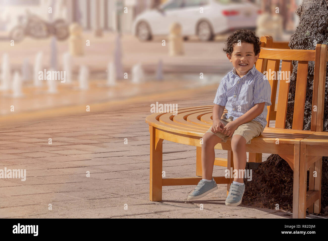 On a sunny day downtown urban city a boy sits on a round bench next to the fountains. Stock Photo