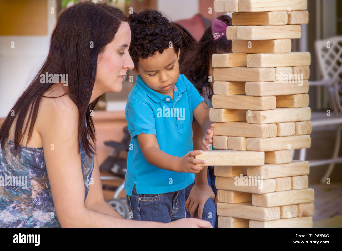 The wooden large size jumbling tower holds steady while the boy keeps it in place with one hand removing a block with the other while Mom is watching. Stock Photo