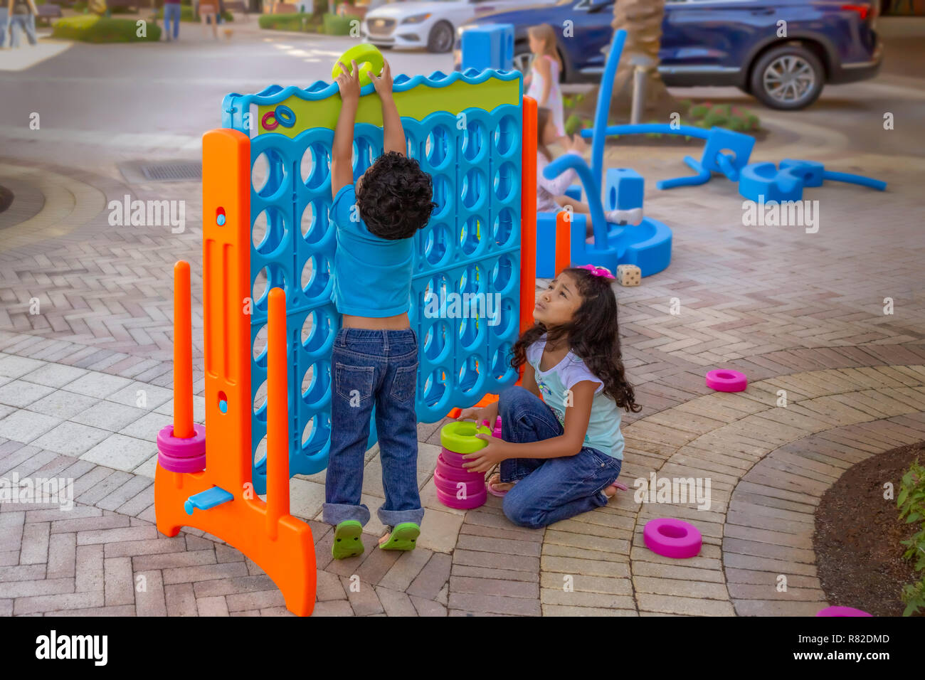 Sister watches younger brother struggle to insert a large ring. Siblings play in a public courtyard with a mega-size connect four-game. Stock Photo