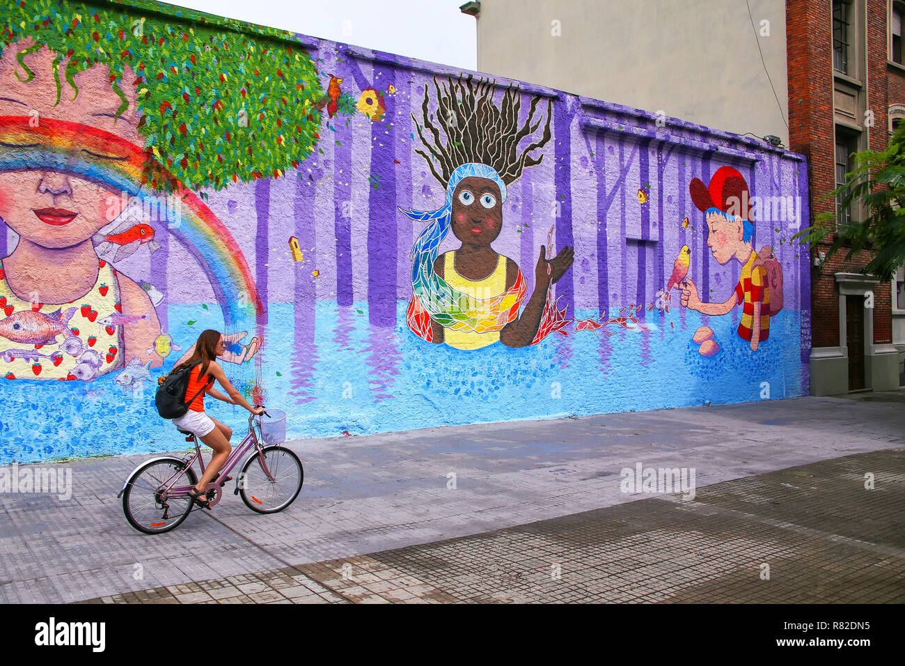 Young woman biking along colorful wall in Montevideo, Uruguay. Montevideo is the capital and largest city of Uruguay. Stock Photo