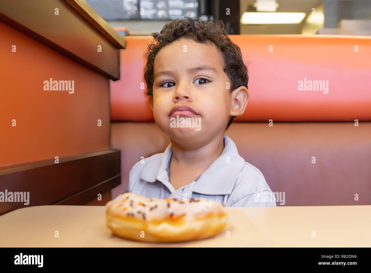 A little boy has a serious expression while he eats a donut in a booth. Head up, eyes focused to the side with a glaring stare. Stock Photo
