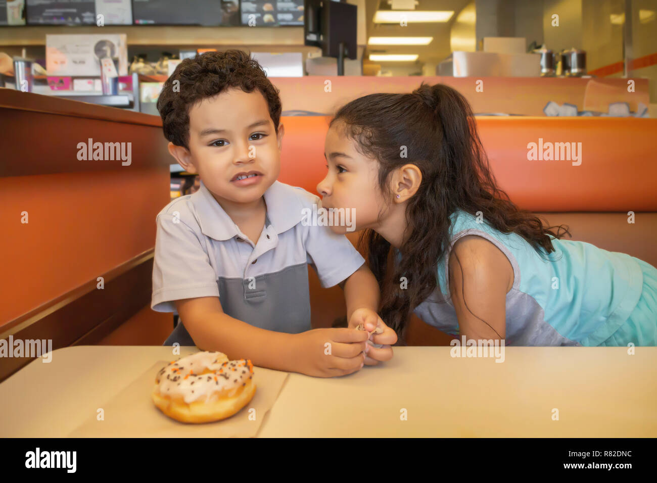 A boy looks at the camera after taking a bite of his donut. Spending time together at a cafe with the older sister is not easy. Stock Photo