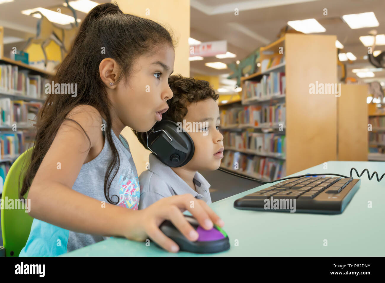 Big sister helps the little brother on the computer at the library. He wears the adult size headphones while she navigates with a wireless mouse. Stock Photo