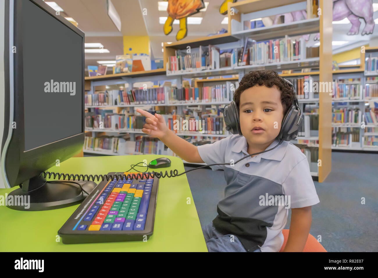 A small boy sits at a desk with headphones pointing at the computer screen. Education at the community public library to learn using the computer. Stock Photo