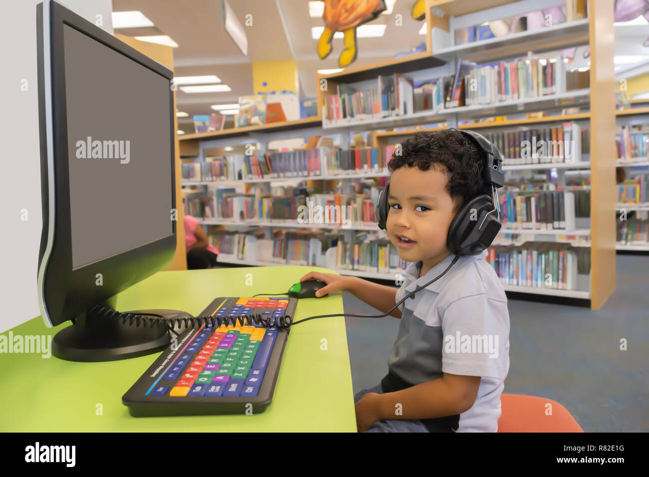 A  fun day at the community public library to play on the computer. The little boy sits at a desk in front of a computer with headphones looking. Stock Photo