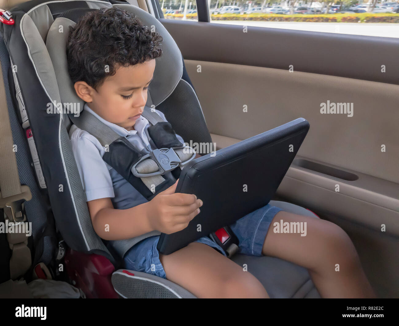 A little boy sits in the car seat playing with a tablet. Focused looking at the tablet pad unaware that he is strapped in the car seat moving. Stock Photo