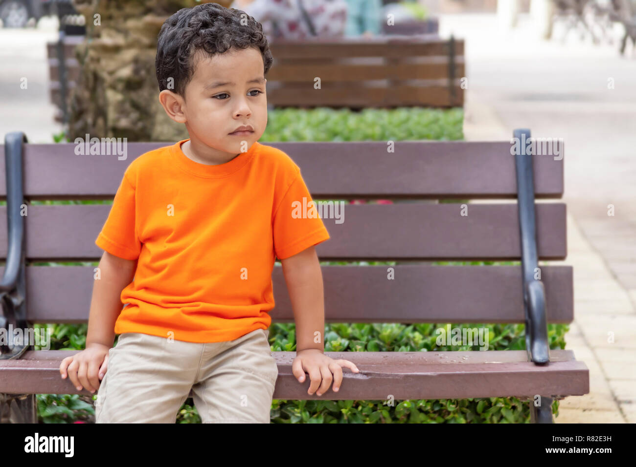 A concerned little boy sits on the public bench. The young boy in the orange shirt sits calmly on the park bench. Stock Photo