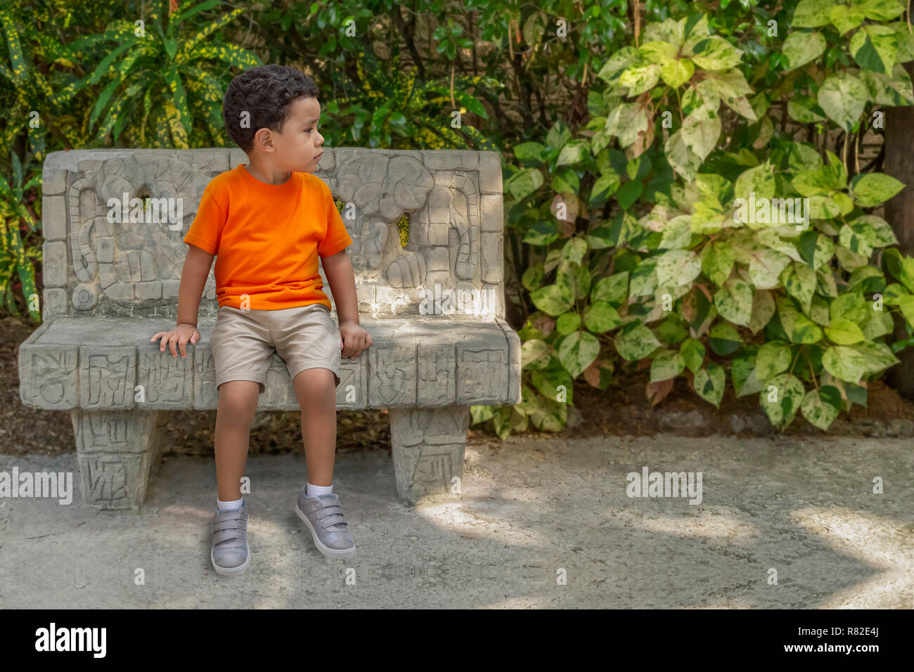 A concerned little boy sits on a cement park bench looking away. He is dressed in, orange t-shirt, tan shorts, and gray shoes sit waiting for mommy Stock Photo