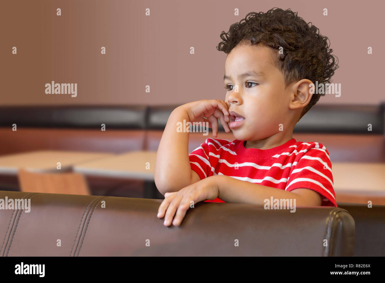 A curly hair young boy looks over the booth seat at a coffee shop. Something at a distance captures the attention of this little boy. Stock Photo