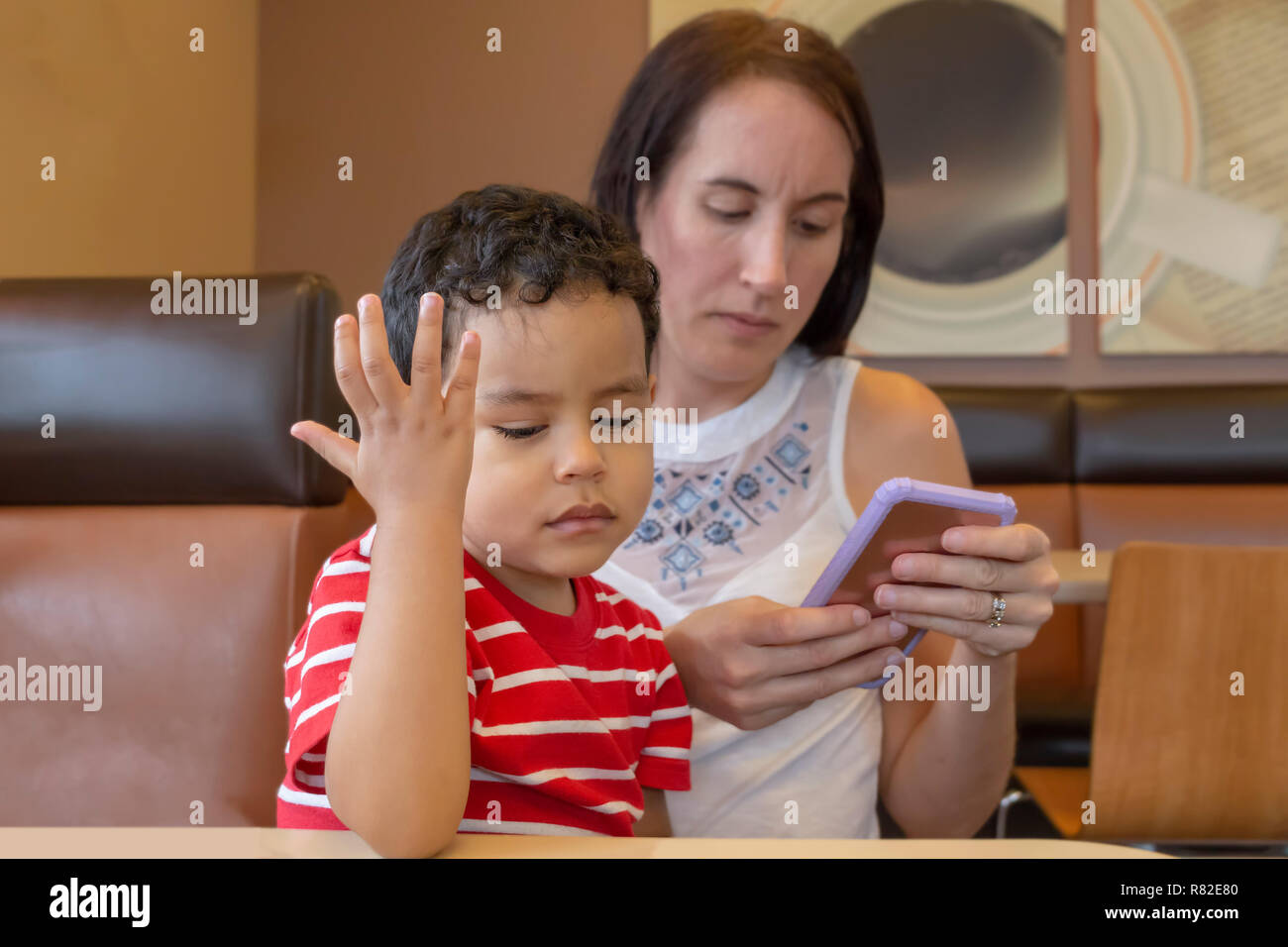 An annoyed little boy is upset at mommy for texting again. A young mother at a coffee shop texting and her little boy is not happy sitting on her lap. Stock Photo