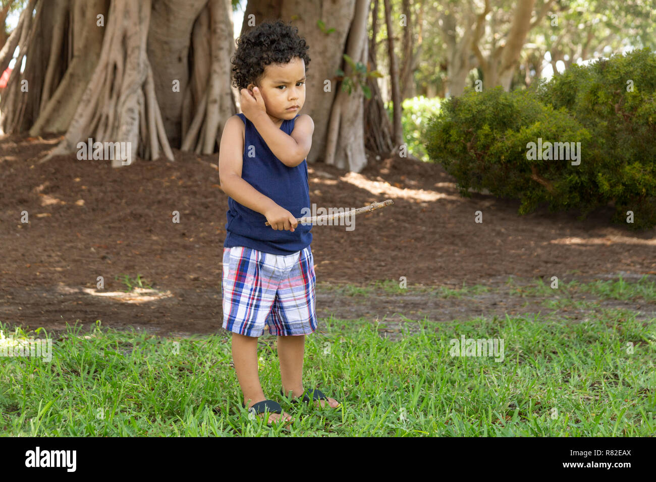 A Hispanic boy with a serious staring look scratches his ear while holding a stick at the community park. Stock Photo