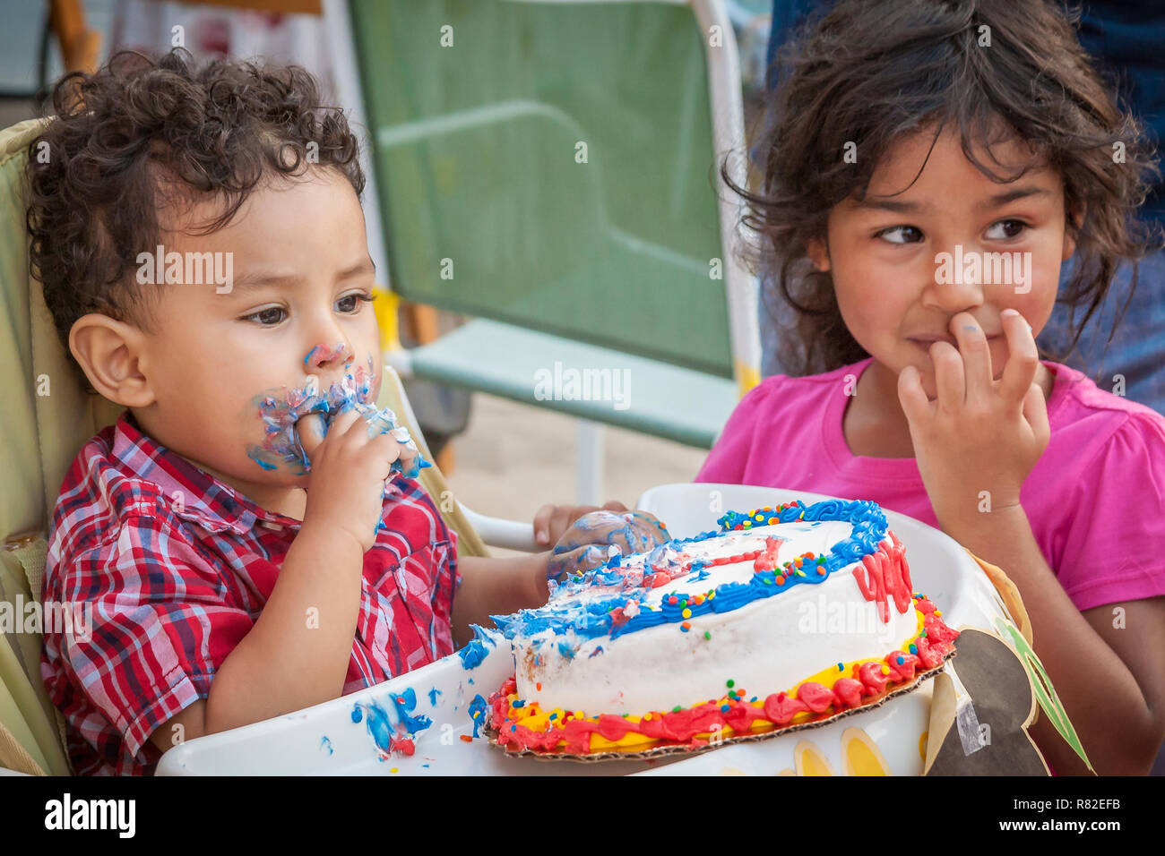 A toddler sets on a baby high chair eating cake with his sister next to him helping. Stock Photo