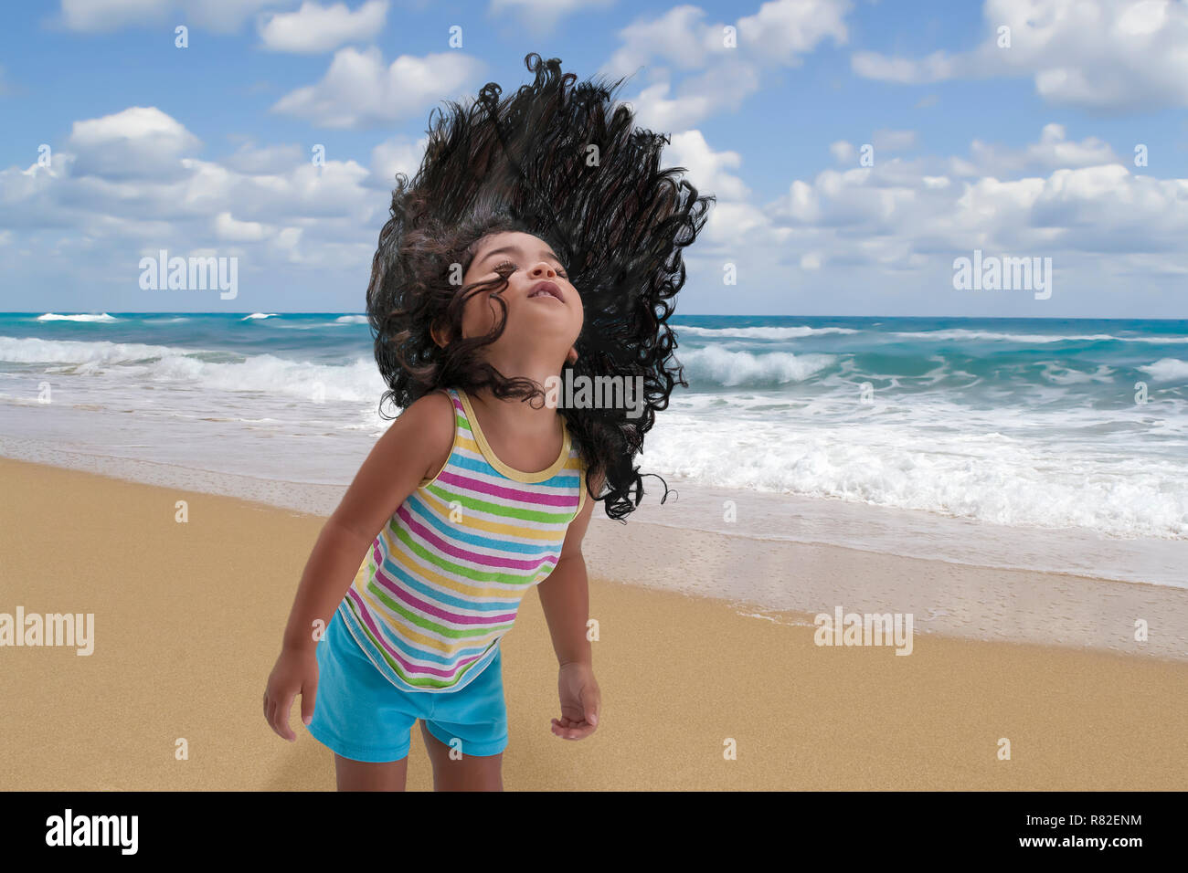 Young girl flings her long black hair upwards with excitement at the beach while the waves crash to the shore in the background. Stock Photo