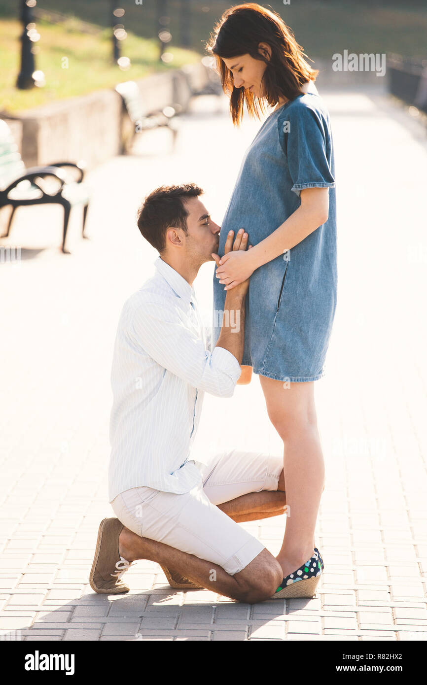 Young man kissing the belly of his pregnant wife while standing on one knee.  A modern couple enjoying a healthy walk together in the park on a sunny d  Stock Photo -