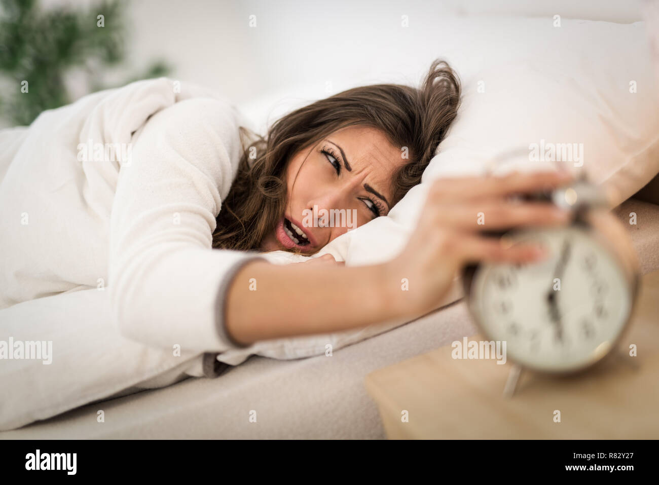 Beautiful young woman frowning in bed and holding hand on alarm clock
