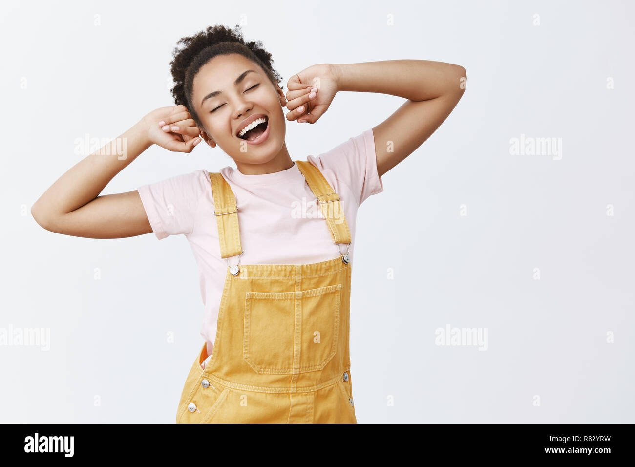 What day to ba awake. Portrait of lazy charming African American female student in stylish yellow overalls, stretching with raised hands, yawning and smiling broadly, feeling sleepy after nap Stock Photo