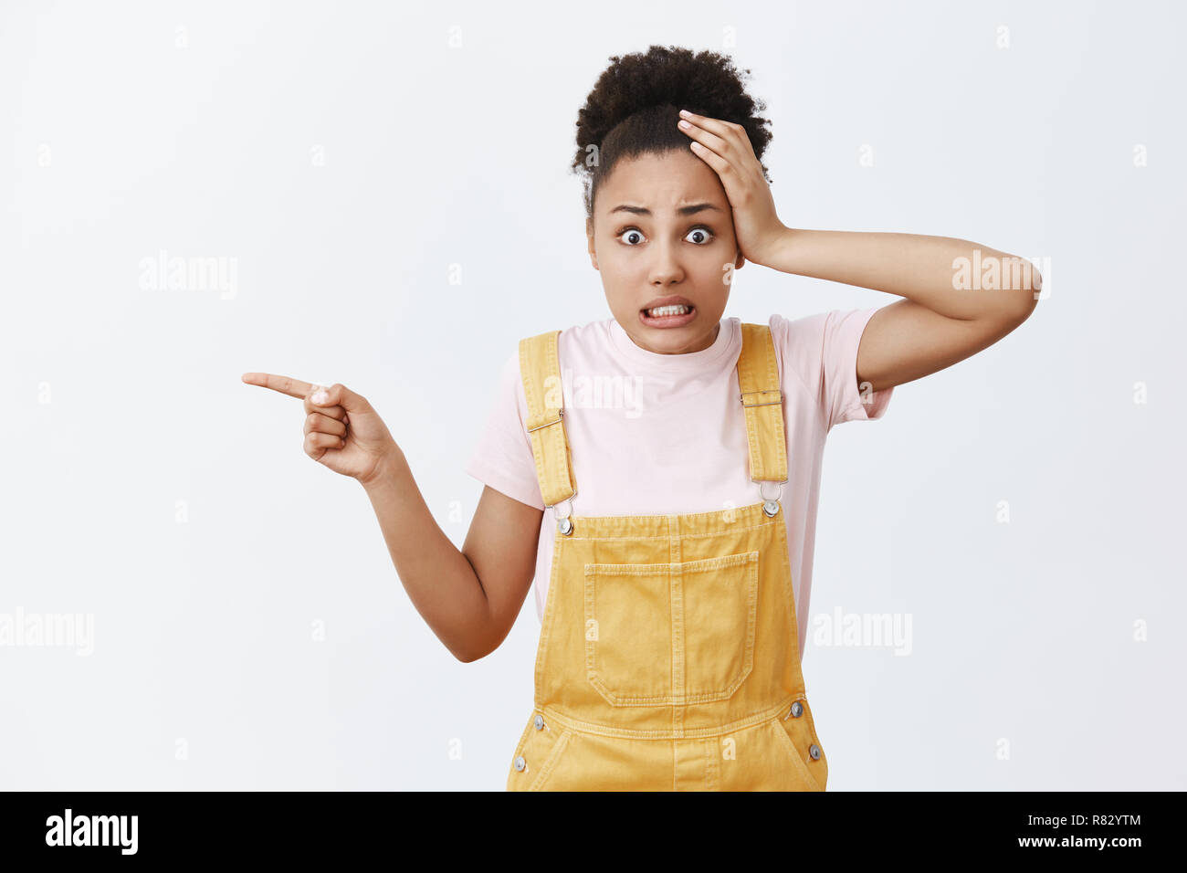 Yikes, what is that. Portrait of shocked worried and confused attractive african-american female with curly hair in fashionable yellow overalls, holding hand on head, pointing left with index finger Stock Photo