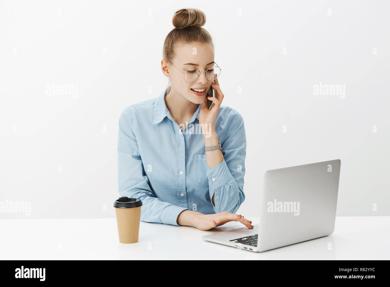 Portrait of charming hardworking female freelancer working with laptop looking at screen, checking order as talking via smartphone with customer, wearing glasses and blue shirt, drinking coffee Stock Photo