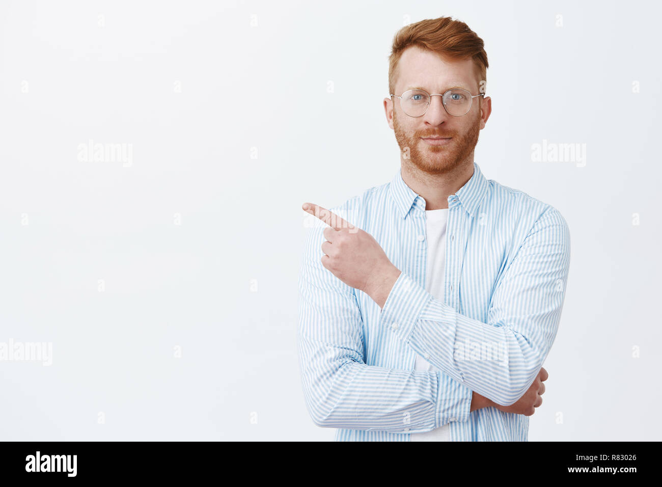 Look there now. Portrait of handsome redhead male with beard in eyewear and shirt pointing at upper left corner and smiling, standing in casual and bossy pose while giving directions to employees Stock Photo