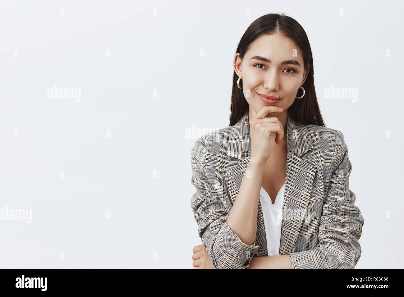 Portrait of charming successful female in stylish jacket over t-shirt, holding hand on chin and smirking while having great idea in mind, thinking about something curious over gray background Stock Photo