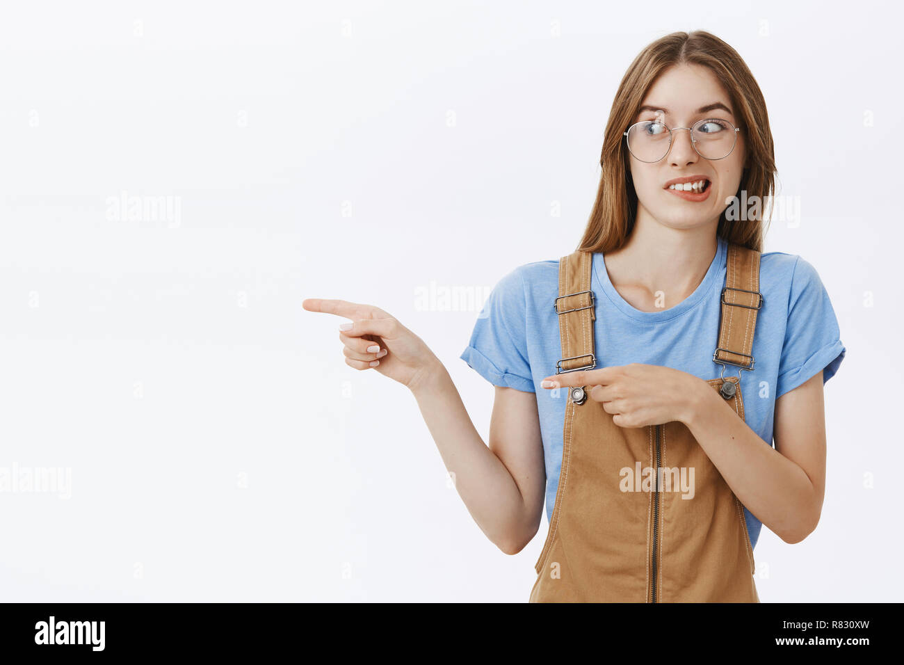 Girl standing with creepy guy showing weirdo to friend. Intense displeased charming european female in brown dungarees and glasses clenching teeth and smirking from antipathy looking and pointing left Stock Photo