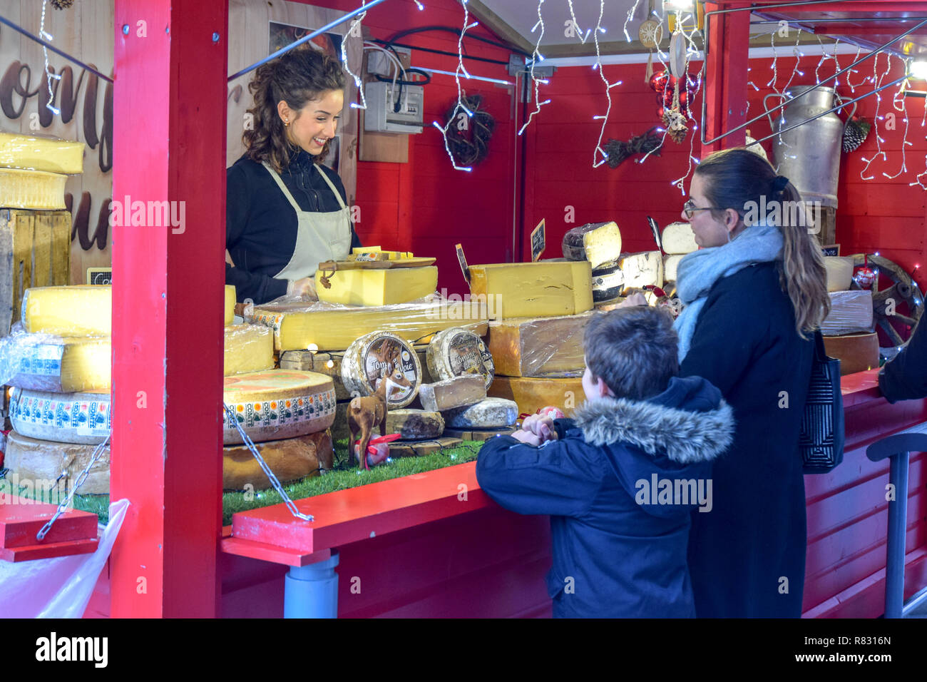 A pretty cheese seller at the Christmas market in Rennes, France waits on a mother and her son. Stock Photo