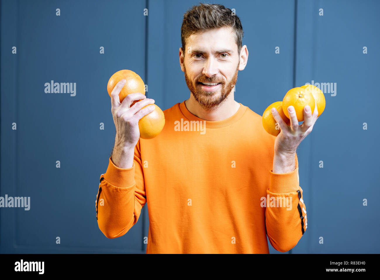 Colorful portrait of a caucasian bearded man in orange sweater holding fruits on the blue background Stock Photo