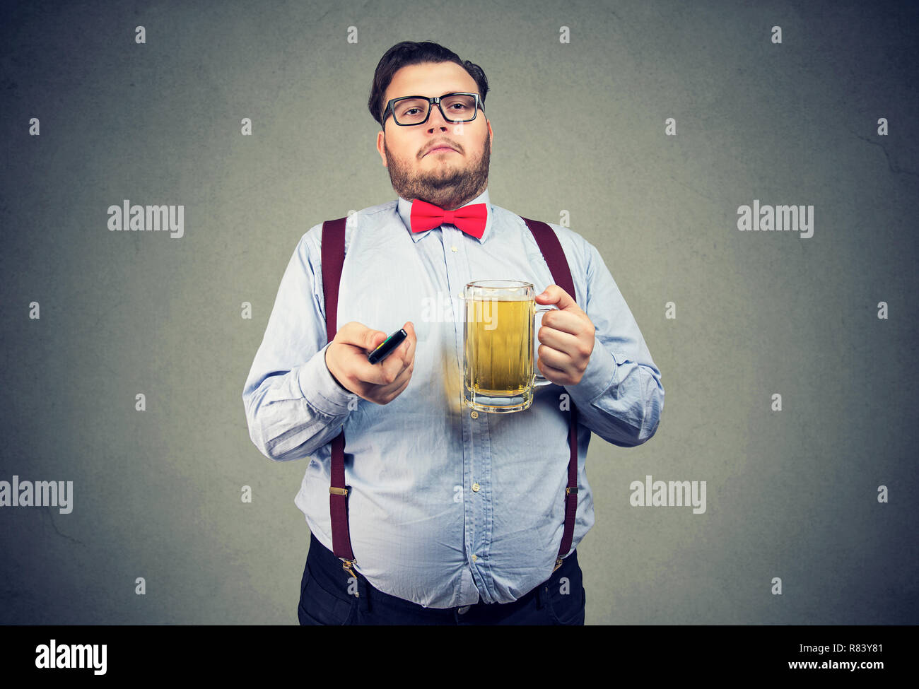 Adult lazy obese man in formal outfit holding mug of beer and watching TV on gray background Stock Photo