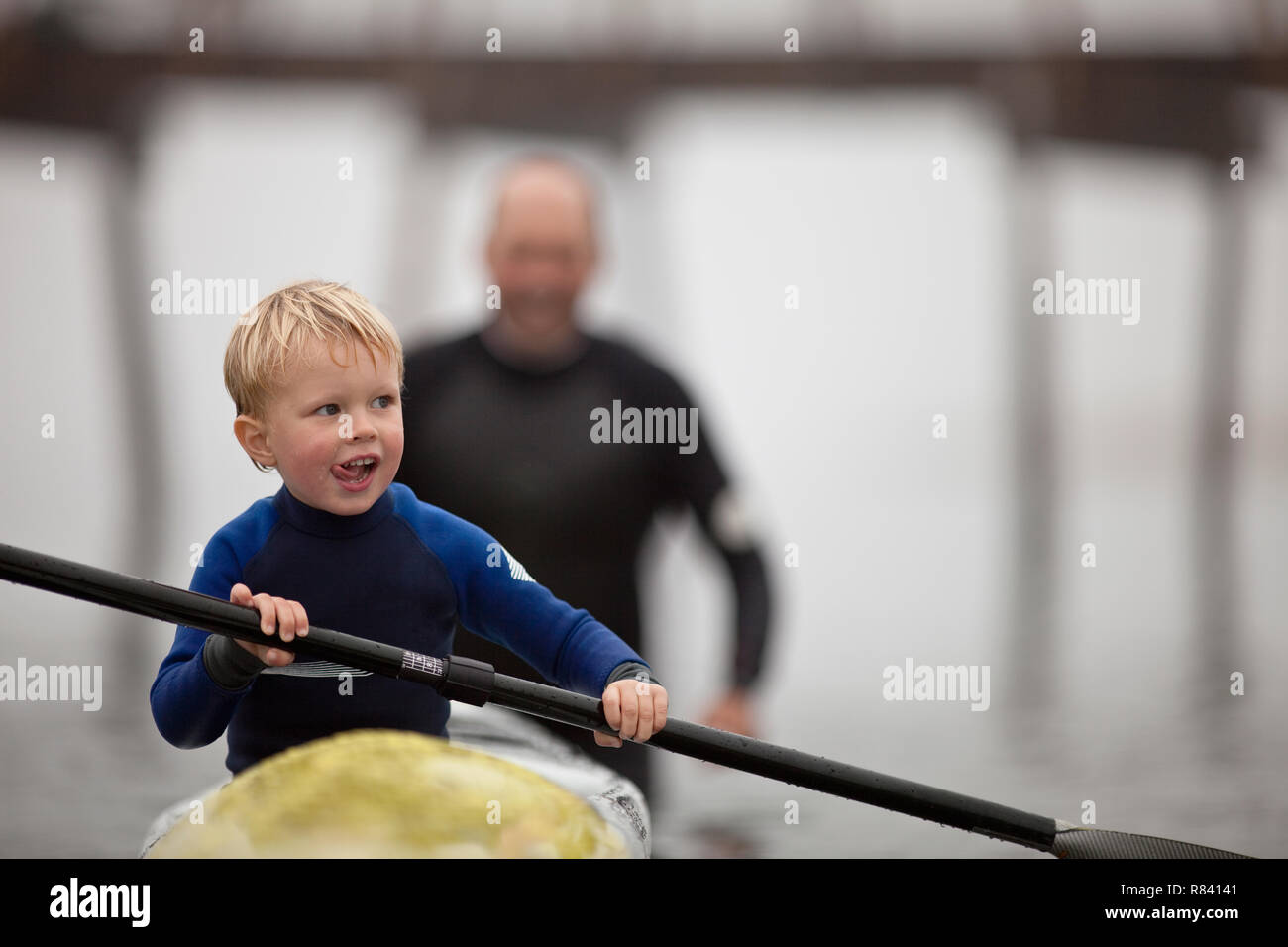Young boy sticks his tongue out in concentration as he tries to row a kayak while his father helps to push it along in the waters of a foggy harbour. Stock Photo