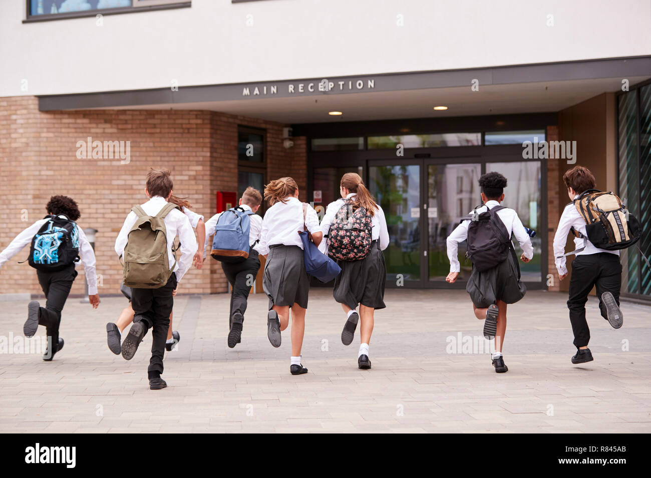 Group Of High School Students Wearing Uniform Running Into School Building At Beginning Of Class Stock Photo