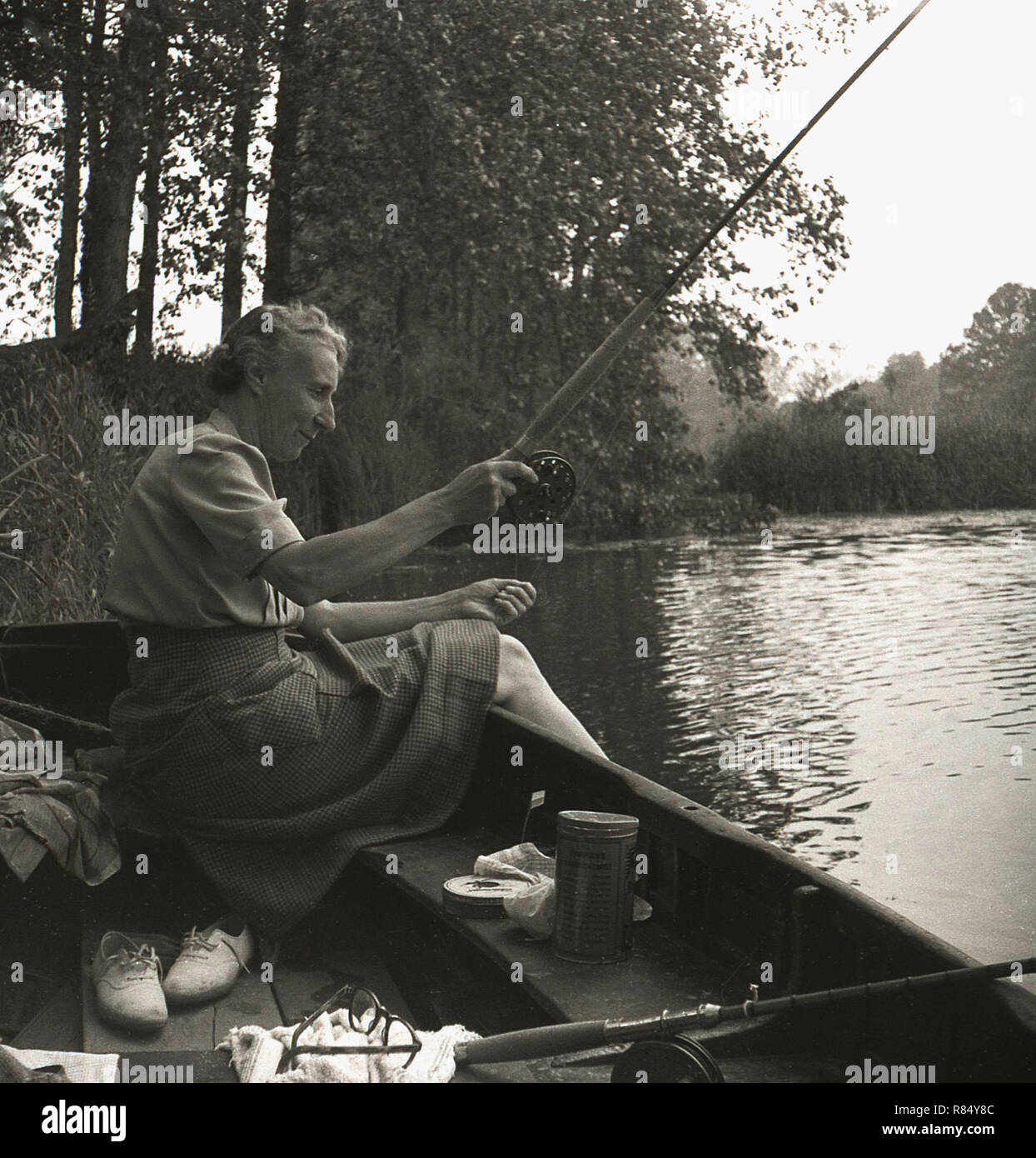 1950s, Elderly lady in a top and skirt sitting in a rowing boat in a river close to the riverbank fishing, England, UK. Stock Photo